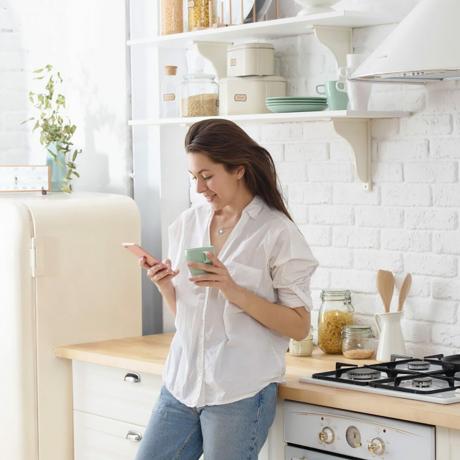 Jonge vrouw met smartphone leunend aan de keukentafel met koffiemok en organizer