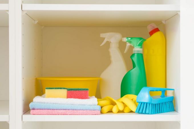 Miska, kefa, fľaše od čistiaceho prostriedku, handry, gumené rukavice a špongie na bielej polici v otvorenej skrini. Zväčšenie. Sada farebných produktov na upratovanie domácnosti. Čelný pohľad.
