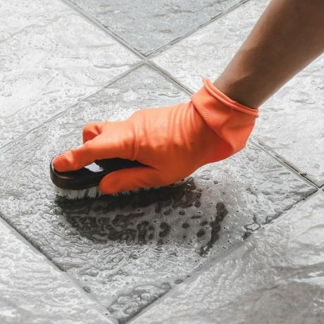 La mano dell'uomo che indossa guanti di gomma arancione viene utilizzata per convertire la pulizia dello scrub sul pavimento di piastrelle.