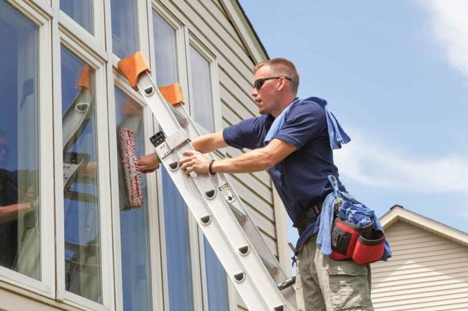 Jauns biznesmenis logu mazgāšanas profesionālis mazgā logus dzīvojamā mājā ar kāpnēm, tīrīšanas sūkļiem un atbilstošu drošības aprīkojumu zilā, saulainā dienā