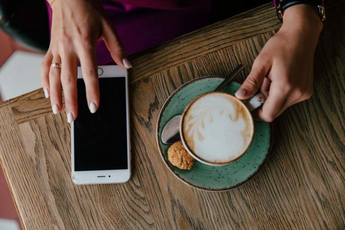 Ženské ruky držia mobilný telefón a šálku cappuccina na tanieri so sušienkou. Na pozadí tabuľky. biela manikúra a telefón. pohľad zhora Čierna obrazovka.