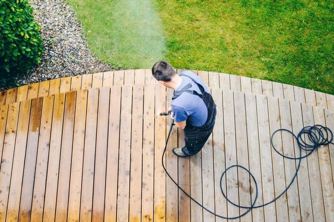 प्रोफेशनल प्रेशर वॉशिंग क्लीनिंग सर्विस मैन लकड़ी की सतह से गंदगी और मलबा हटाने के लिए पावर वॉशर के साथ छत के डेक या आँगन पर उच्च दबाव वाले पानी का छिड़काव करता है।