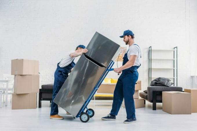 двое грузчиков в униформе на ручной тележке перевозят холодильник в квартире
