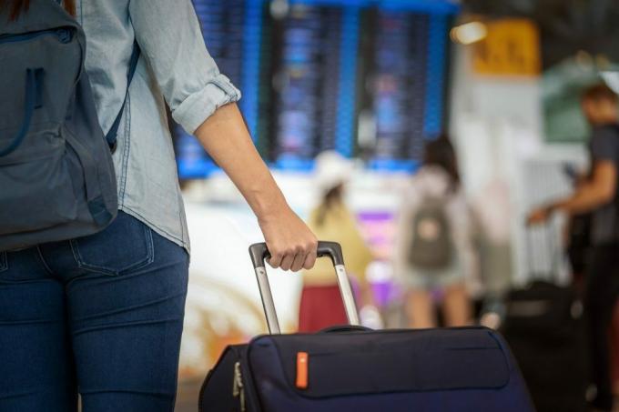 יד תקריב שמחזיקה את המזוודות מעל לוח הטיסה לצורך צ'ק-אין במסך פרטי הטיסה בתכנון מודרני של שדה תעופה, נסיעות ותחבורה.