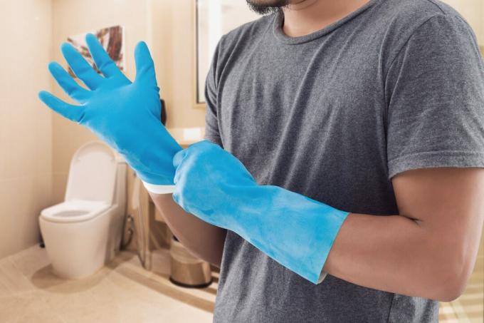 Vīrietis pelēkā kreklā, kas valkā zilus gumijas cimdus pirms tualetes mazgāšanas.