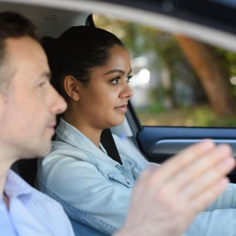 Teini-ikäinen tyttö saa ohjeita ajaessaan autoa mieheltä matkustajan istuimella