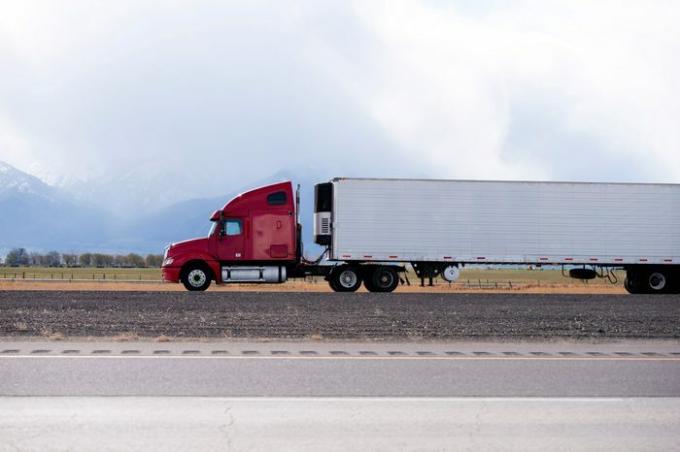 Oldalnézetből a világos piros nagy rig félig teherautó -flotta, amely rakományt szállít hosszú utaló félpótkocsiban a lapos úton Utah -ban, hóval borított hegyekkel és felhőkkel a háttérben