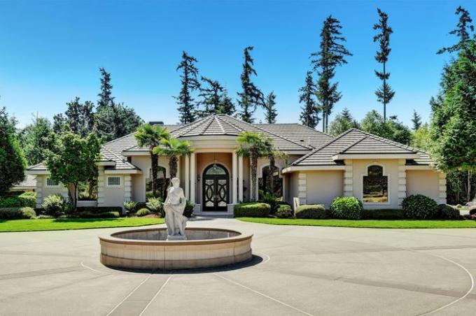 Casa unifamiliar suburbana con estatua de fuente en el patio delantero, entrada de asfalto. Casa residencial de lujo con grandes ventanales, árboles alrededor y fondo de cielo azul. Noroeste, EE. UU.