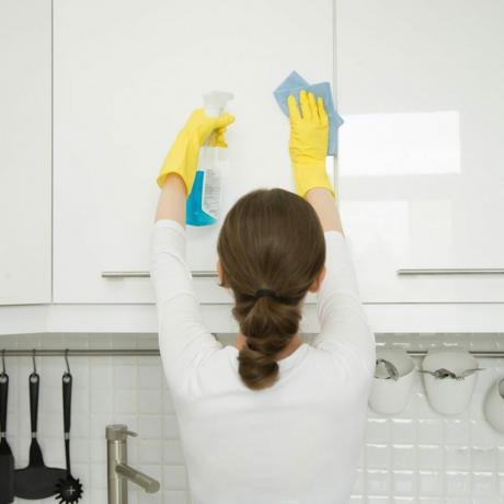 Bakifrån på en attraktiv ung kvinna som rengör en yta av det vita köksväggskåpet, bär gummi skyddande gula handskar, med trasa och sprayflaskmedel.