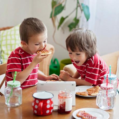 Duas crianças felizes, dois irmãos, tomando café da manhã saudável, sentados à mesa de madeira na cozinha ensolarada, comendo waffles e assistindo desenhos animados no tablet