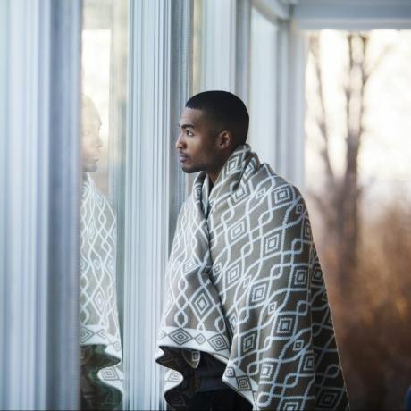 Vyras, uždengtas antklode, žiūri į šalį stovėdamas prie lango namuose