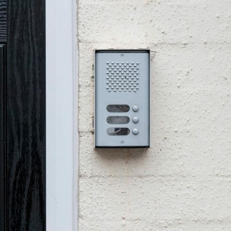 stanovanjsko-zvočniški-domofon-poleg-črnih-vrat-na-beli steni