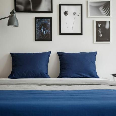 포스터가 있는 흰색 침실 내부의 테이블과 램프 사이의 침대에 네이비 블루 베개가 있습니다. 실제 사진