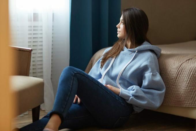 Inomhusskott av tankeväckande mörkhårig kvinna klädd i blå tröja, jeans, sitter på golvet nära sängen, tittar eftertänksamt på fönstret, poserar i mysigt sovrum i modern lägenhet. Inhemsk atmosfär