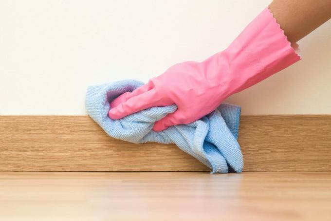 Запослени руку у гуменој заштитној рукавици са крпом од микро влакана брише подножје на поду од прашине са зида. Пролећно генерално или редовно чишћење. Концепт компаније за комерцијално чишћење.