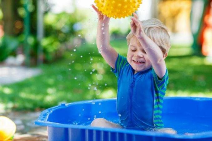 Kis totyogó fiú szórakozik a fröccsenő vízzel és labdázik a nyári kerti medencében