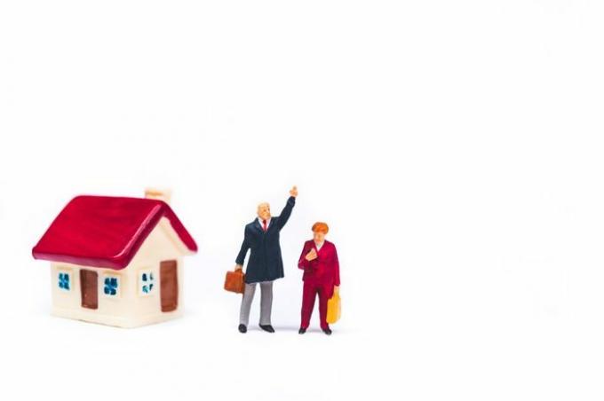 Miniatūri cilvēki, vīrietis un sieviete, kas stāv pie mini mājas izolētā uz balta fona, izmantojot biznesa un ģimenes koncepciju