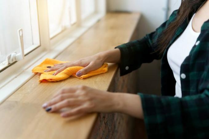 Žena v domácnosti upratuje drevený pult vedľa okna a odstraňuje prach utieraním oranžovej handry. Šťastný koncept čistenia.