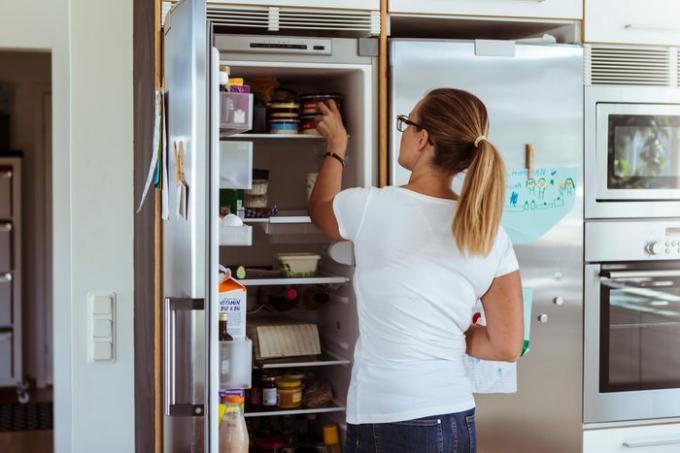 Skats no aizmugures uz sievieti, kas skatās ledusskapī, stāvot virtuvē