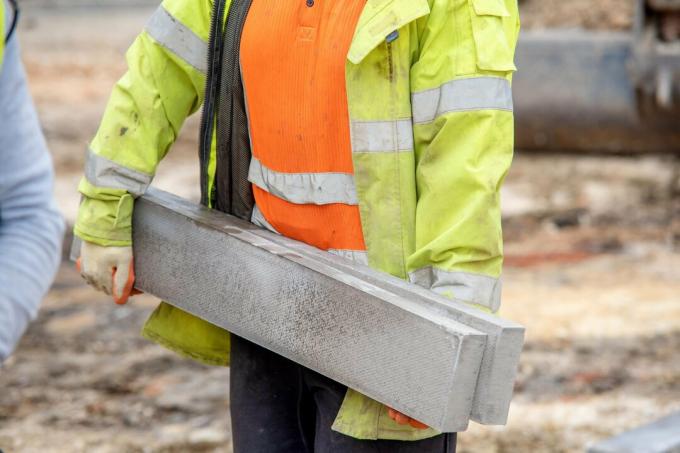 Bližnji posnetek zemeljskega delavca v oranžnem in rumenem hi-vizu, ki nosi težke betonske robnike na gradbišču med gradnjo nove ceste