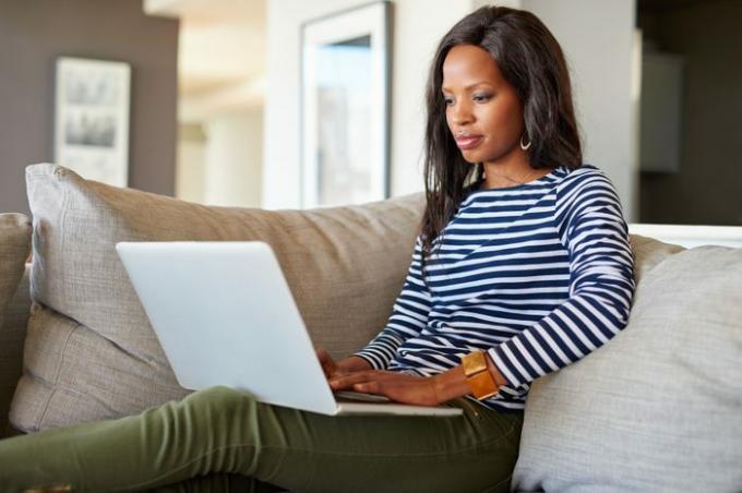 आकर्षक युवती घर में सोफे पर आराम करते हुए लैपटॉप का उपयोग करती है