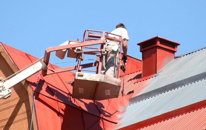 عامل تاجر يقوم بتشغيل رافعة ويطلي سقفًا معدنيًا لمنزل باللون الأحمر باستخدام بخاخ طلاء في يوم أزرق مشرق