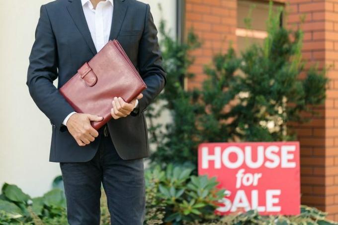 Primer plano del empresario en traje negro con maletín de cuero marrón de pie junto a una casa en venta