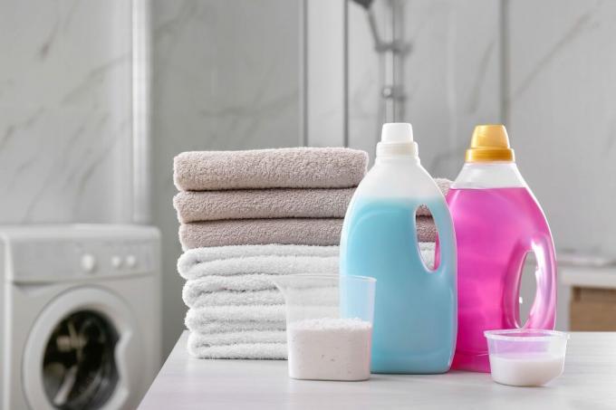 Pila de toallas plegadas y detergentes sobre mesa blanca y lavadora en segundo plano.