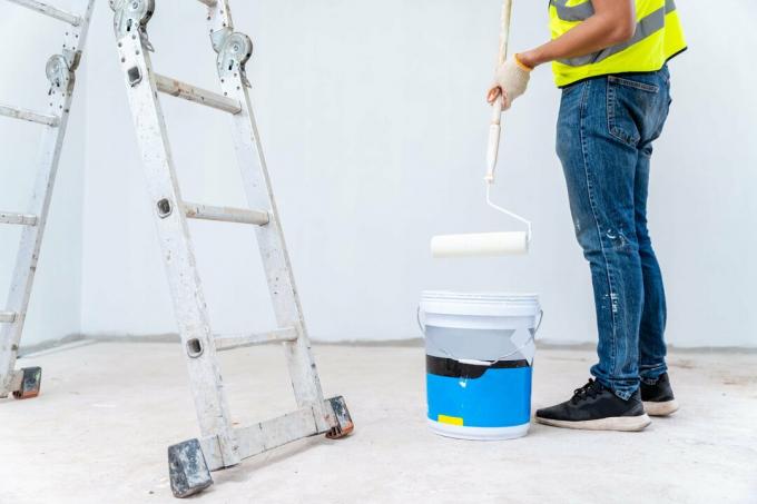 Pintor pintando la pared, con rodillo de pintura y balde en una casa sin terminar, trabajo de construcción, aislado en un gran espacio vacío con escalera